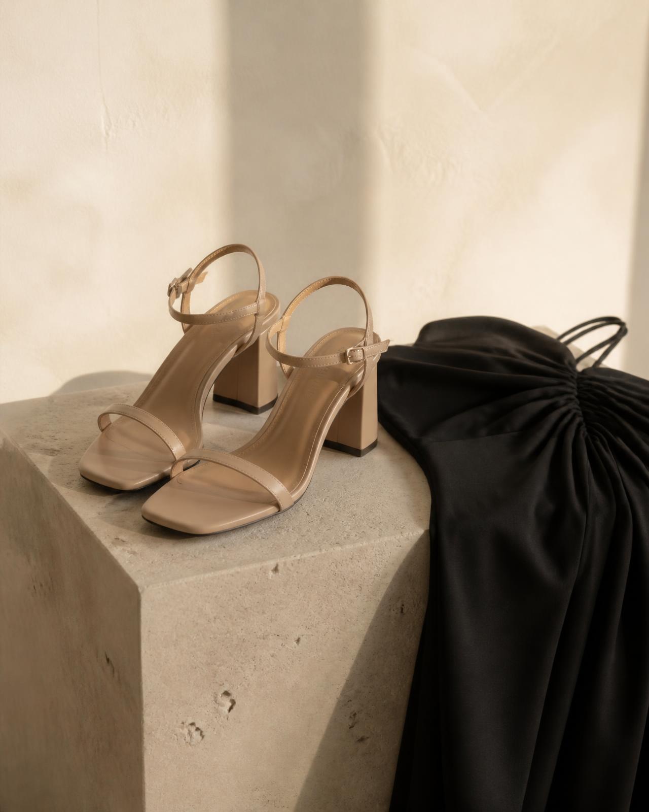 Nude block heel sandals styled beside a black satin dress on a stone plinth